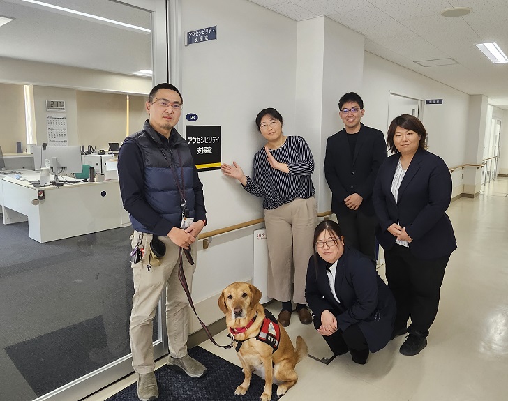 支援室前での集合写真。左から愛媛大学太田さんと聴導犬のハンナさん。中央後列　左からスタッフの萩原・岡田・吉田が立ち、前に磯田がしゃがんでいる。